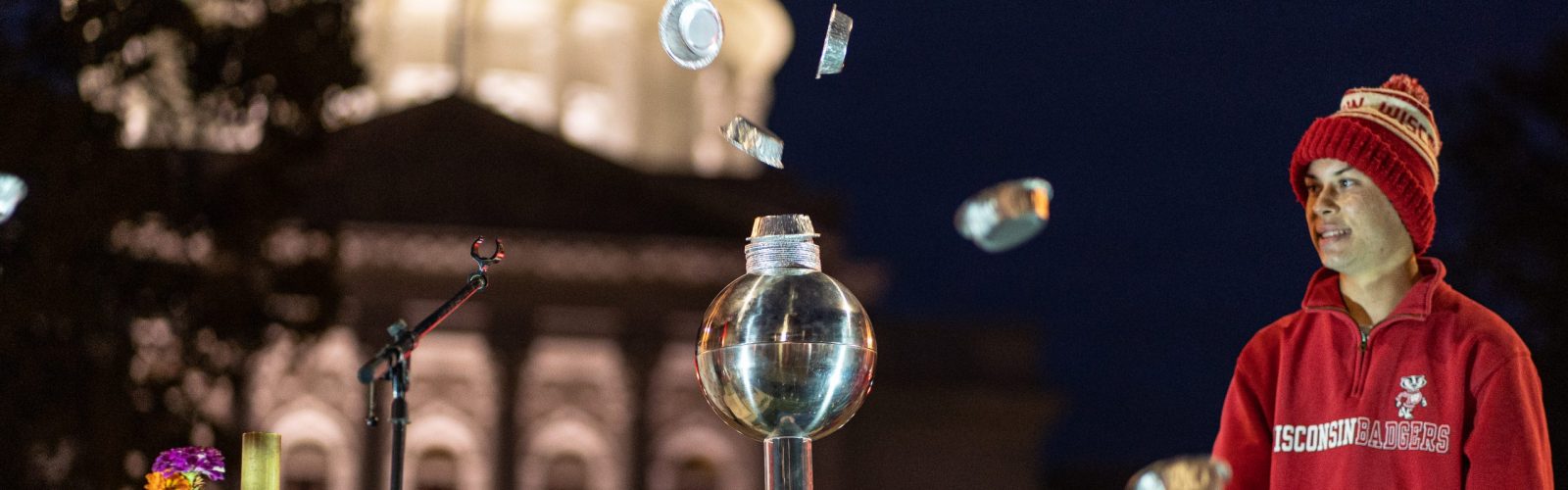 a man wearing a Wisconsin sweatshirt and hat stands in front of the illuminated State Capitol building at dusk, with a Van der Graff machine to his right and aluminum pie plates floating in the air