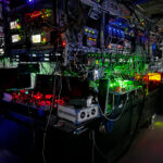 a photograph of a room with the lights off, but the bulk of the image is taken up by a large piece of complicated equipment with many different colored laser lights visible, illuminating the shape of the equipment
