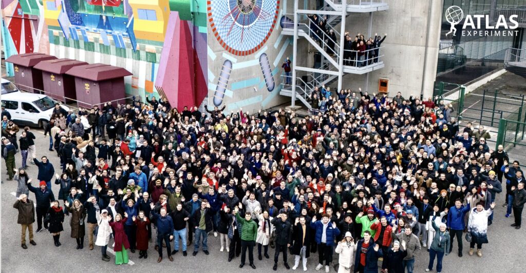 thousands of people stand as a group in front of some vaguely science-y looking (and very large!) equipment