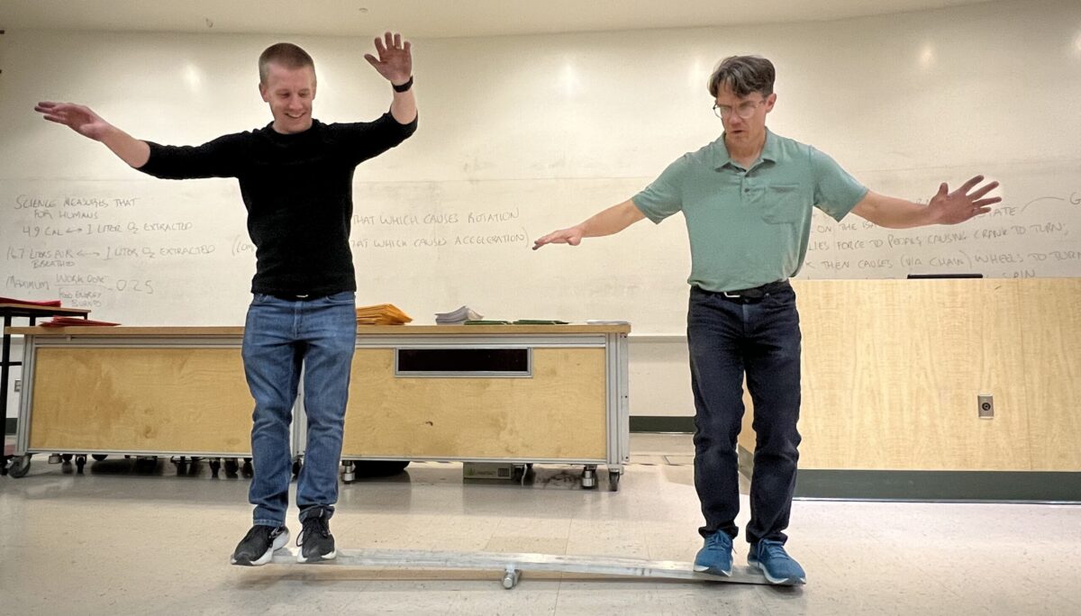 two people stand and balance on either side of a plank with a fulcrum under it. both their arms are out like they are trying not to fall.